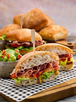 Food photograph of a ham salad sandwich, a golden sourdough sandwich roll filled with curled folded slices of cured ham, thick slices of fresh tomato, shredded lettuce and mustard. Served on parchment paper on a wooden board with bowls of lettuce salad and other sandwiches, with a second board piled high with sandwich rolls shot on a grey background