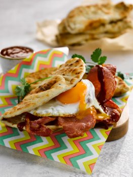 Food photograph of a bacon and egg naan, a golden folded naan bread filled with crispy fried bacon and a fried egg with a runny yolk which is pouring over the bacon underneath, served on brightly patterened baking paper, on a light grey tabletop with extra naan bread in the background