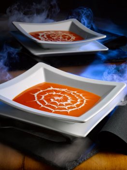 Food photograph of a Hallowe'en soup, two square white bowls of tomato soup with a swirl of double cream made into a spider web pattern, on a dark background with blue light illuminating wisps of smoke