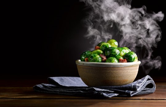Food photograph of an artisan ceramic bowl of fresh bright green steamed brussels sprouts with crispy bacon lardons and chestnuts, served on a denim tea towel on a dark wooden tabletop, shot against a black background with steam billowing from the hot brussels sprouts