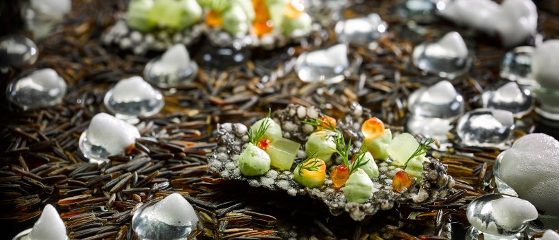 Food photograph of a fine dining canape, a squid ink tapioca cracker topped with cubes of cucumber jelly, avocado puree, salmon caviar and dill leaves. Shot on a reflective chrome background covered with wild rice, glass pebbles and foam