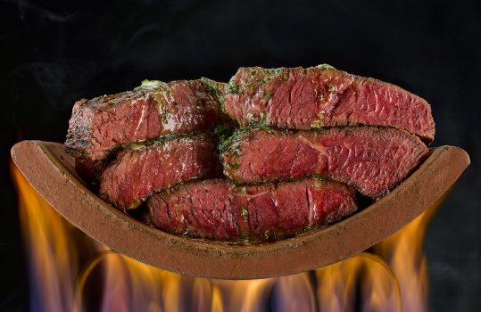 Abstract food photograph of five beef rib eye steaks, seared and cooked rare, stacked on a terracotta tile and topped with melting garlic and parsley butter - shot on a black background with wisps of smoke, and flames underneath the tile