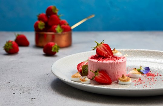 Food photograph of a fine dining dessert, strawberry parfait with freeze dried strawberry powder, fresh strawberries, burnt marshmallow and edible flowers, served on a white plate on a grey stone table with a copper pot full of fresh strawberries against a bright blue background