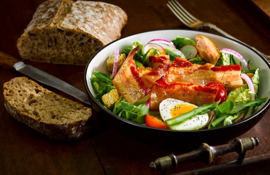 Food photograph of a bacon and egg salad, soft boiled egg and crispy bacon tossed with crispy croutons, sliced red onion, sliced tomato, mixed leaves and shaved asparagus, shot on a vintage dark wooden serving tray alongside a loaf of homemade brown bread