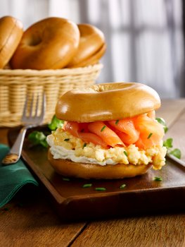 Food photograph of a smoked salmon and scrambled egg bagel, a shiny golden toasted bagel topped with a layer of cream cheese, fluffy scrambled egg sprinkled with chives, and slices of glistening smoked salmon, served on a dark wooden board in a home setting, a wooden tabletop with a basket of bagels, and a window with net curtains in the background