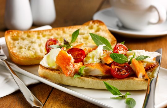 Food photograph of a crispy golden toasted ciabatta topped with flaked deep brown smoked trout, along with charred cherry tomatoes, torn mozzarella and sprigs of basil - shot in a hotel style setting on a white plate with white salt and pepper shakers, and a white teacup and saucer on a wooden tabletop