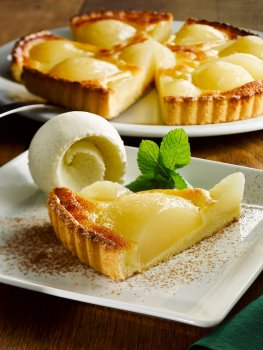 Food photograph of a slice of tarte aux poires, crisp short pastry crust filled with halved pears and frangipane, served on a square white plate with a swirled scoop of vanilla ice cream, shot on a wooden table with the whole tart in the background on a large plate