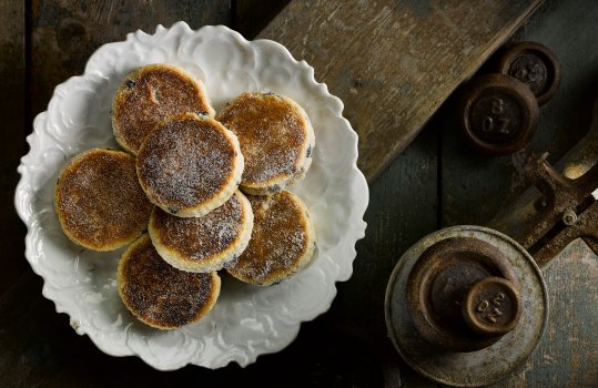 Aerial food photograph of a vintage plate of homemade traditional Welsh cakes, golden brown Welsh cakes dusted with sugar, shot on a weathered wooden background with vintage cast iron baking weights and weighing scale