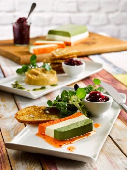 Food photograph of a three layer tricolour vegetable terrine, sliced and served on a white rectangular plate with mixed leaves, sweet chilli sauce, chutney and melba toast - shot on a multi coloured weathered wooden background