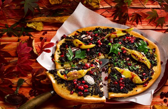 Aerial food photograph of a Turkish style topped flatbread, crispy baked flatbread base topped with spiced fried minced beef, pomegranate seeds, pickled chillies, picked parsley leaves and pomegranate mollases drizzle. Served on parchment paper with a vintage bread knife lifting a slice of flatbread, on a red painted wooden background with scattered autumnal leaves
