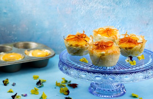 Food photograph of homemade orange and vanilla custard tarts, crisp layers of filo pastry filled with vanilla custard and a slice of orange, and topped with candied orange peel. Four of these tarts served on a vintage blue glass cake stand on a bright blue backdrop, with scattered spring flowers, and a muffin tin with more tarts in the background