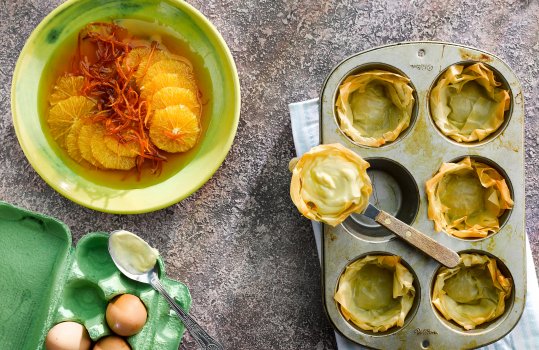 Aerial food photograph showing the making of vanilla custard and orange filo pastry tarts, six crispy filo shells in a muffin tin, one having been removed and filled with custard, shot alongside a box of eggs, and a bowl of orange slices and candied orange peel, marinating in syrup, shot on a grey stone background