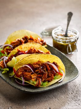 Food photograph of vegan pulled jackfruit tacos, crispy corn tortilla taco shells filled with pulled barbecue jackfruit, sliced red onion, lettuce and sliced red pepper, served on a grey platter alongside a jar of homemade salsa
