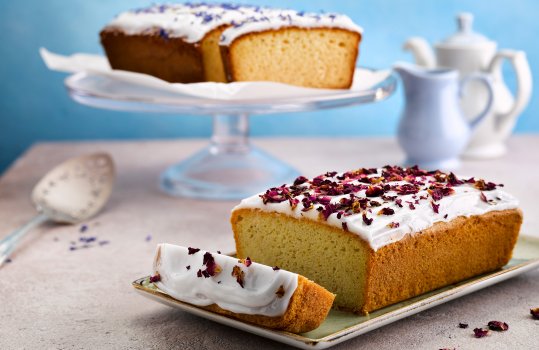 Food photograph of a pair of vegan loaf cakes, in the foreground a cardamom and rose loaf topped with white icing and dried rose petals is served sliced on a rectangular plate, with dried rose scattered around, and in the background a lemon loaf topped with icing and dried bergamot flowers is served sliced on a glass cake stand alongside a vintage cake slice, and a teapot and milk jug - all shot on a grey stone table with a bright blue backdrop