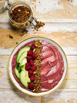 Aerial food photograph of a vegan smoothie bowl, a thick bright pink mixed berry smoothie flecked with dark purple from the skins, topped with quinoa and cashew granola, fresh raspberries and fresh avocado. Served in a white china bowl on a weathered wooden background, alongside a jar of the homemade quinoa and cashew granola
