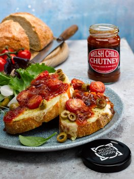 Food photograph of a cheese and tomato toast topped with Welsh chilli chutney, thick slices of sourdough toast topped with grilled melted cheese, charred cherry tomatoes and olives, and then topped with Welsh chilli chutney and served with mixed salad leaves and pickled onions - shot on a light grey stone table with a loaf of sourdough bread in the background alongside a jar of Welsh chilli chutney, in front of a bright blue backdrop