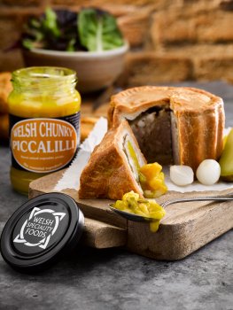 Food photograph of a large sharing size pork pie sliced and served on parchment paper on a vintage wooden board with pickled silverskin onions and Welsh chunky piccalilli - shot in a home style setting on a dark grey tabletop with a bowl of mixed salad leaves and a jar of Welsh chunky piccalilli, with a red brick wall in the background