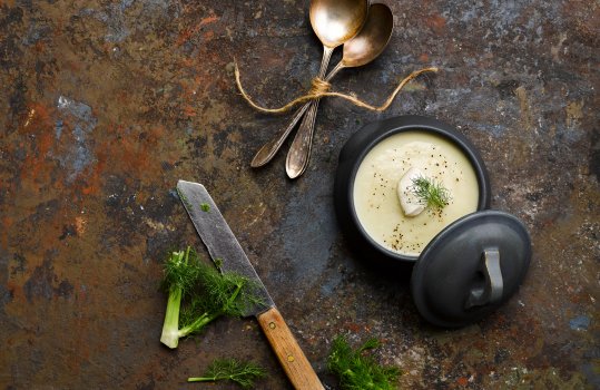 Aerial food photograph of homemade fennel and apple soup, topped with fennel fronds, cracked pepper and creme fraiche, served in a ceramic bowl with a lid offset to one side - shot on an abstract painted dark background alongside a vintage chefs knife, chopped fennell tops and a bundle of vintage soup spoons