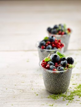Food photograph of vanilla and lime chia seed puddings, served in plastic takeaway pots and topped with fresh blueberries, pomegranate seeds, sprigs of mint and lime zest - three of these pots shot on a white wooden tabletop with grated lime zest scattered around