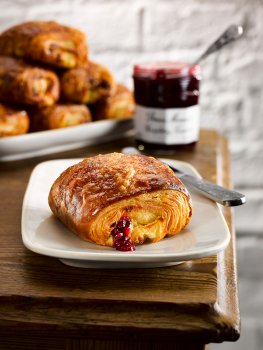 Food photograph of a golden crisp flaky raspberry croissant served on a white plate on the corner of a farmhouse table, with a platter of croissants in the background alongside a jar of raspberry jam