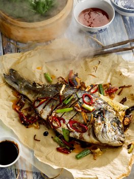 Aerial food photograph of a whole baked Asian style sea bream, whole sea bream baked in parchment paper with soy sauce, chilli, spring onion, ginger and garlic - shot on a painted blue background with a steam basket of cabbage leaves, and small dishes of sweet chilli, and hoisin sauces