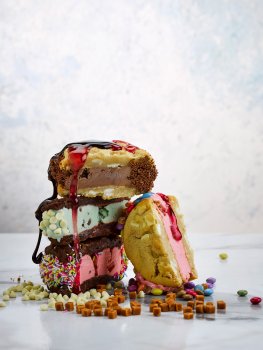 Food photograph of a selection of ice cream sandwiches, various flavours of ice cream sandwiched between chocolate and chocolate chip cookies, and then rolled in either sprinkles, smarties or white chocolate chips and sliced in half - a stack of these sandwiches drizzled with strawberry and chocolate sauces and served with scattered smarties, fudge pieces and white chocolate chips on a marble table in front of a pale blue background