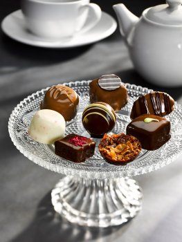 Food photograph of a selection of premium dark, milk and white chocolates on a glass cake stand, with a white teapot, cup and saucer in the background