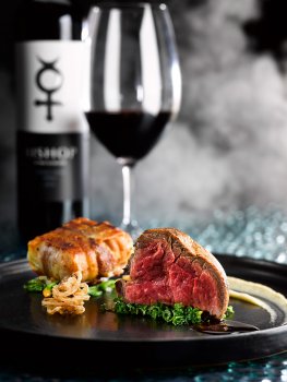 Food photograph of a juicy glistening rare beef fillet steak, garnished with crispy onions and horseradish puree, and served alongside a crispy bacon wrapped dauphinoise potato. Served on a black plate on a glass background with a glass and bottle of red wine, and clouds of smoke