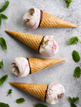 Aerial food photograph of four chocolate waffle ice cream cones topped with scoops of blackberry ripple ice cream and shot on a grey stone background with scattered mint leaves