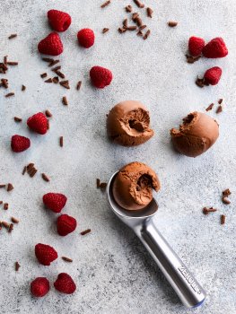 Aerial food photograph of three curled scoops of vegan chocolate ice cream, shot on a light grey stone background with scattered raspberries and chocolate curls