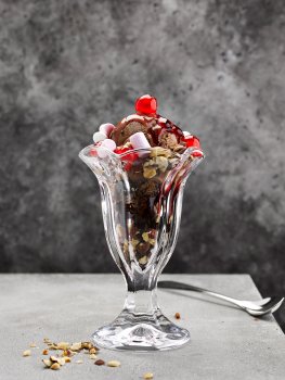 Food photograph of an ice cream sundae featuring Belgian chocolate ice cream, chunks of chocolate brownie, mini marshmallows, chopped almonds, strawberry syrup and glace cherries, served in a tall sundae glass with scattered chopped almonds on a grey table in front of a dark background
