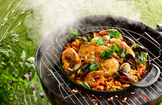 Food photograph of a barbecue chicken, king prawn, mussel and clam paella, a cast iron pan shot on a lit barbecue filled with a paella of cooked rice, clams and mussels in their shells, large king prawns and chicken thighs with seared crispy skin, sprinkled with parsley and show outdoors with foliage and billowing smoke