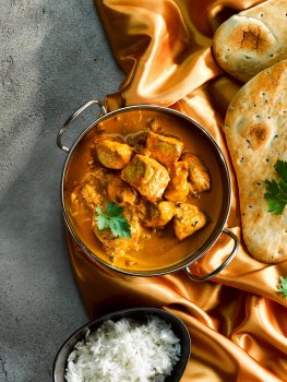 Aerial food photograph of a chicken tikka masala with large chunks of chicken coated in shiny orange sauce, served in a balti dish alongside a bowl of steamed rice and golden naan bread - shot on shimmering orange fabric on top of a grey stone background