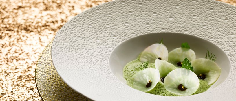 Food photograph of a fine dining starter, cones made from raw kohlrabi, with laver seaweed inside, in a foaming seaweed broth - served in a large white decorative bowl on top of a matching gold plate, shot on a glittering gold background