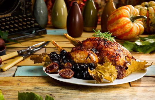 Food photograph of a whole roast chicken with dark shiny glazed skin, served with roasted figs and black grapes on a white plate on a rustic wooden background with seasonal squash, scattered autumn leaves, artisan ceramic vases and an antique typewriter
