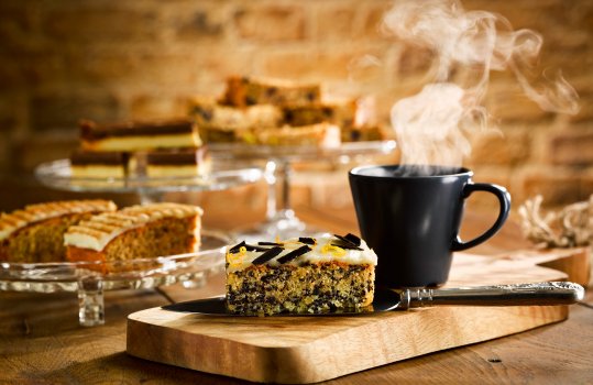 Food photograph of a chocolate and vanilla traybake slice with thick shiny icing, served on a cake slice on a wooden board in a cafe setting - with glass cake stands of other cake slices in the background, alongside a steaming cup of coffee