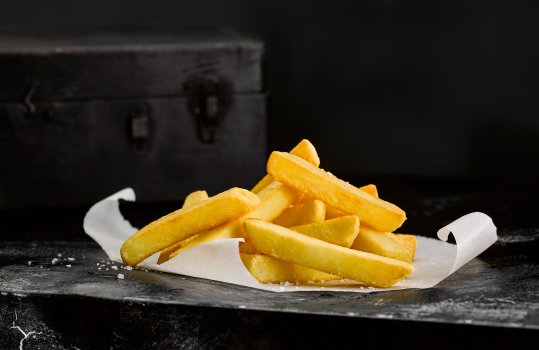 Food photograph of a pile of golden crispy thick cut chips, sprinkled with large flakes of sea salt served on parchment paper, on a black background with a black granite tabletop and a black wooden chest in the background