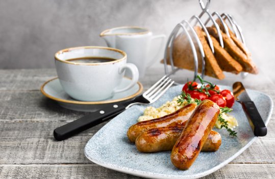Food photograph of a homemade breakfast, three thick pork sausages with shiny brown skins, served on a pale blue plate alongside scrambled eggs and roasted vine cherry tomatoes, shot in a home style setting with brown toast in a vintage toast rack, alongside a cup of black coffee and a milk jug, on a grey background