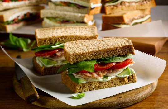 Food photograph of a takeaway meal deal BLT sandwich, slices of streaky bacon with thick sliced fresh tomato and lettuce leaves between slices of thick cut brown bread served on parchment paper on a wooden board - shot on a dark wood set with other filled sandwiches in the background
