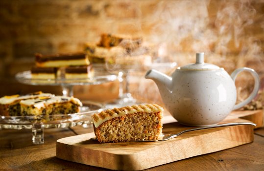 Food photograph of a carrot cake traybake slice with thick shiny icing, served on a cake slice on a wooden board in a cafe setting - with glass cake stands of other cake slices in the background, alongside a steaming teapot