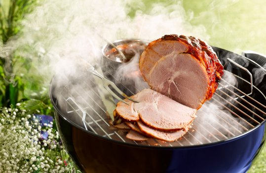 Food photograph of a glazed barbecue ham, a whole ham joint with a shiny sticky brown glazed exterior and slices being taken from the front, shot in an outdoor setting on a lit barbecue alongside a saucepan of glaze, with foliage in the background and smoke clouds billowing from the barbecue