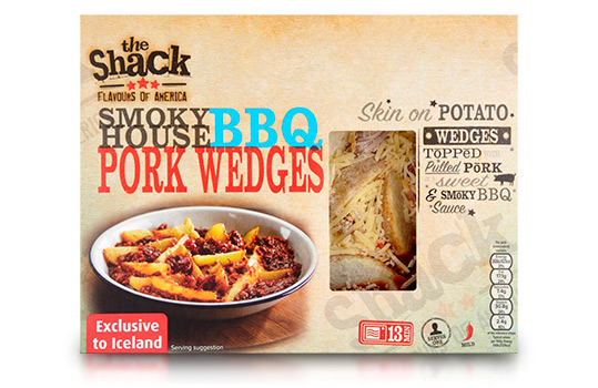 Food photograph of a front of pack image for The Shack Smoky BBQ Pork Wedges frozen ready meal, an enamel dish filled with shiny BBQ pulled pork with potato wedges and sliced jalapeno, shot on a dark wood setting with ingredients in the background, and the front of pack itself shown over the photograph