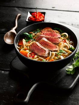 Food photograph of a Pan-Asian noodle broth, rice noodles with carrot, spinach, chilli and coriander topped with a seared medium rare beef sirloin steak cut into slices, shot on a dark set in a black bowl on a black wooden board on a black wooden table, with a black dish of chopped chillies and a dark wood carved spoon