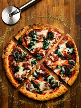 Aerial food photograph of an artisan sourdough pizza base topped with tomato sauce, spinach, sundried tomatoes and mozzarella cheese, sliced into four quarters and sprinkled with dried oregano which is also scattered on the dark wood background