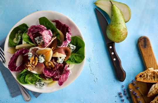 Aerial food photograph of a pear, walnut and roquefort salad, a poached pear along with leaves of radicchio, lettuce and chicory, topped with toasted walnuts and crumbled roquefort cheese. Shot on a bright blue stone background alongside a fresh halved pear and golden toasted French bread