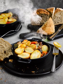 Food photograph of a lamb hot pot frozen ready meal, chunks of braised lamb and vegetables topped with crispy sliced potato served in a cast iron dish on a black plate, served alongside large chunks of crusty brown bread, shot on a light grey stone setting with billowing clouds of steam