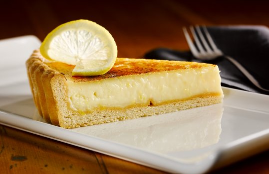Close up food photograph of a slice of tarte au citron, short crumbly thin pastry filled with a smooth thick lemon custard, and topped with blowtorched caramelised sugar, garnished with a slice of lemon and served on a white rectangular plate on a dark wooden table