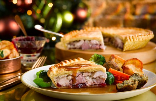Food photograph of a Christmas dinner pie, crisp flaky golden pastry filled with large chunks of turkey and ham with cranberry sauce, topped with gravy and served with crispy golden roast potatoes, carrots, brussels sprouts and stuffing balls. Served on a white plate and shot on a gold reflective tabletop with the whole pie on a board in the background next to a bowl of cranberry sauce; with a Christmas tree and a red brick wall in the background