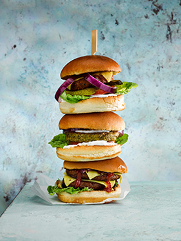 Food photograph of a stack of vegan burgers, various vegan patties topped with lettuce, cheese, red onion, pickles, ketchup and mayonnause between vegan style brioche buns, served in a stack threaded onto a bamboo skewer and shot in an abstract sky blue setting