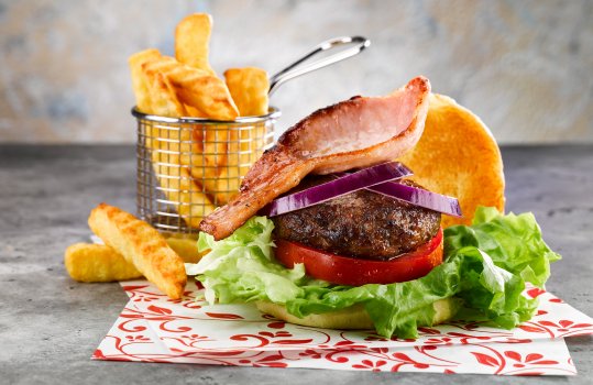 Food photograph of a homemade burger and chips, a golden toasted bun filled with large lettuce leaves, thick sliced fresh tomato, a dark crusted juicy burger patty, sliced red onion and a rasher of crispy fried bacon served on parchment paper alongside a basket of crinkle cut chips on an abstract grey background